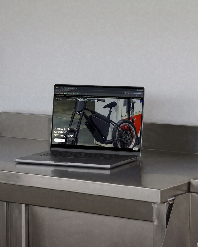A laptop on a stainless steel bench showing the F1 Ebikes site, its hero reading A NEW ERA OF RIDING STARTS HERE.